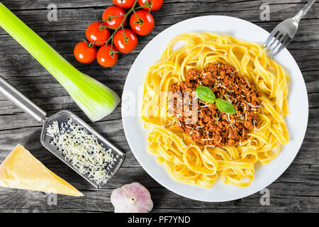 Bolognese Ragout mit italienischer Pasta auf einer weißen Platte, dekoriert mit Basilikumblätter, authentische Rezept, Holz- Hintergrund mit Sellerie, Knoblauch, Cherry tom Stockfoto