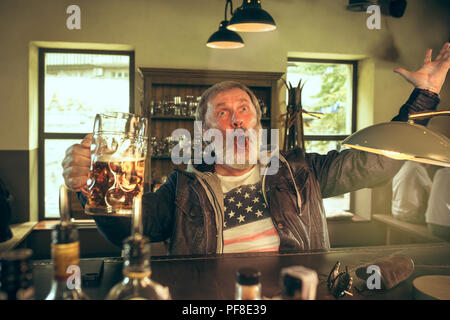 Älterer bärtiger Mann Alkohol trinken im Pub und einen Sport Sendung im TV. Mein Favorit wimmelt und Bier genießen. Mann mit Becher Bier am Tisch sitzen. Fußball oder Sport Fan. Menschliche Emotionen Konzept Stockfoto