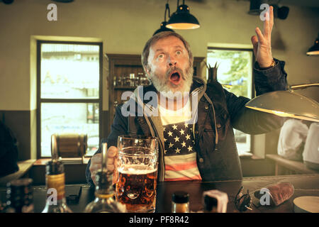 Älterer bärtiger Mann Alkohol trinken im Pub und einen Sport Sendung im TV. Mein Favorit wimmelt und Bier genießen. Mann mit Becher Bier am Tisch sitzen. Fußball oder Sport Fan. Menschliche Emotionen Konzept Stockfoto