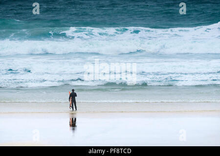 Ein einsamer Surfer, der mit seinem Surfbrett an der Küste auf den Fistral Beach in Cornwall. Stockfoto