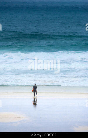 Ein einsamer Surfer, der mit seinem Surfbrett an der Küste auf den Fistral Beach in Cornwall. Stockfoto