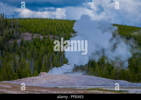 Schönen blick auf Old Faithful Geyser Basin im Yellowstone National Park gelegen, durch Dampf mit einem grünen Berg hinter im wunderschönen sonnigen Tag umgeben Stockfoto