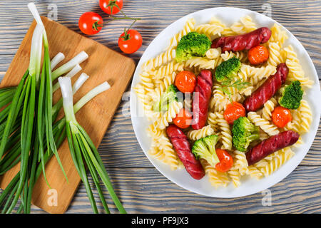 Köstliche Spirale Nudelsalat mit Brokkoli, gegrillte Würstchen und Tomaten auf einem Teller, Frühlingszwiebeln auf einem Schneidebrett, Ansicht von oben Stockfoto