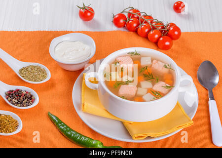 Lachs Suppe mit Karotte und Kartoffel, gewürzt mit Dill in Weiß Schüssel serviert auf einem orange Serviette, auf einem weißen Holztisch, mit Sahne und Gewürzen, Stockfoto