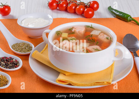 Lachs Suppe mit Karotte und Kartoffel, gewürzt mit Dill in Weiß Schüssel serviert auf einem orange Serviette, auf einem weißen Holztisch, mit Sahne und Gewürzen, Stockfoto