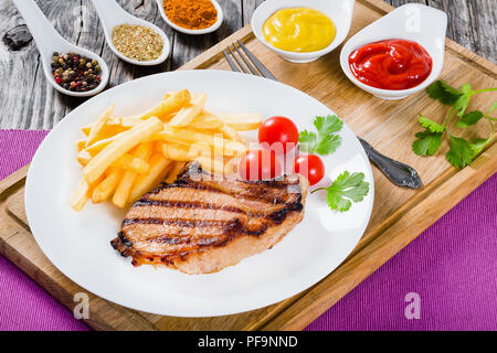 Gegrillte Schweinekoteletts auf einem weißen Teller mit Pommes Frites, Tomaten, Koriander Blätter und Gewürze, auf einem Holztisch, close-up Stockfoto