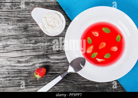 Gekühlt Erdbeeresuppe mit Stücken von Erdbeeren und Minze in einem weißen breiten Felge dish auf einem Tisch Matte, Creme in einer Soße Boot auf einem alten rustikalen t Stockfoto