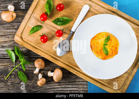 Französische Küche - Julienne Gratin in Schale mit Blättern auf weissem Teller mit Löffel dekoriert, auf alten Mainboards mit Basilikum, Tomaten und Pilze auf backgrou Stockfoto