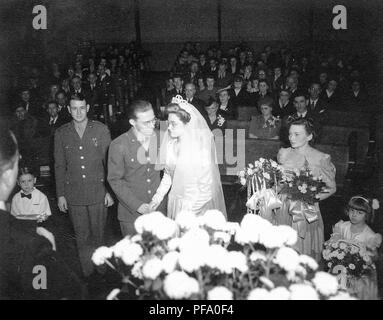 Schwarz-weiß Foto, geschossen von hinter der rechten Schulter der Ehe Dienstleiter und zeigt ein Bräutigam in eine militärische Uniform, neben einem Braut trug ein weißes Kleid, Schleier, Tiara, und Gläser, mit dem besten Mann (auch in Uniform) eine junge Ring bearer, das Mädchen der Ehre, mit Blumen, einem jungen Brautjungfer mit Blumen, und eine große Anzahl von Gästen in den Kirchenbänken im Hintergrund sitzt, in Ohio, 1945 fotografiert. () Stockfoto