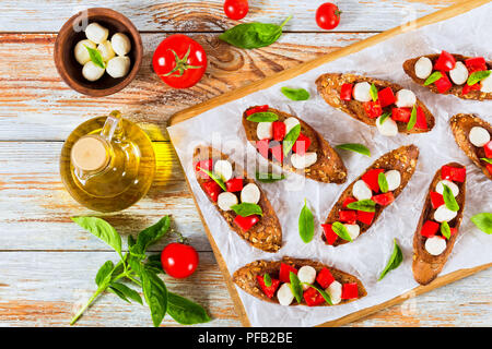 Bruschetta mit Tomaten, Mozzarella und Basilikum auf Olivenöl Baguette mit Samen, auf Schneidebrett auf Pergamentpapier serviert gebraten, Bo Stockfoto