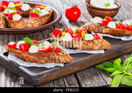 Bruschetta mit Tomaten, Mozzarella und Basilikum auf Roggen Baguette mit Samen, auf Pergamentpapier auf dunklen Holz- Board. Und auf Lehm Teller, italienisches Rezept, Cl Stockfoto