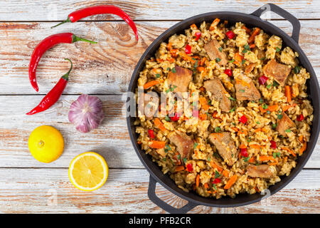 Hausgemachte zubereitete Paella mit Fleisch, Pfeffer, Gemüse und Gewürze in eiserne Pfanne auf weißem abblätternde Farbe holzbohlen, Chili, Zitronenscheiben und Ga Stockfoto