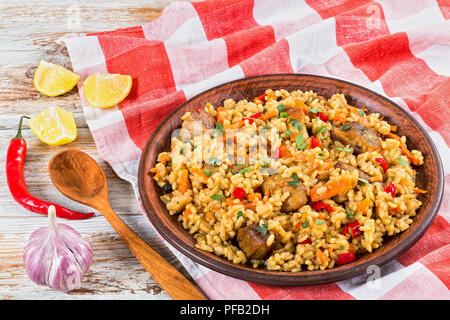 Reis mit Fleisch, Pfeffer, Gemüse und Gewürzen auf Ton Schüssel, Küche Handtuch, Zitronenscheibe, Knoblauch, Chili und Holzlöffel auf weißem abblätternde Farbe pl Stockfoto