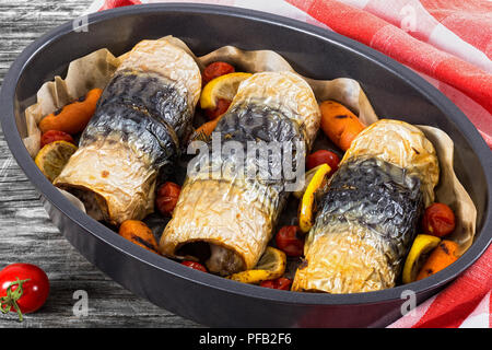 Gesunde gebackenes Filet Makrelen mit Karotten, Kirschtomaten und Zitrone in oval Auflaufform, auf dunklen Holzbrettern mit Küchentuch, Ansicht von Abo Stockfoto