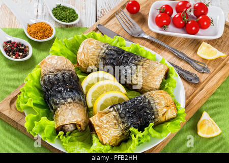 Gesunde gebackenes Filet Makrelen in Rollen auf Salatblättern und Zitronenscheiben auf weißem Teller auf Schneidebrett mit Messer und Gabel, Gewürze und Platte mit Stockfoto