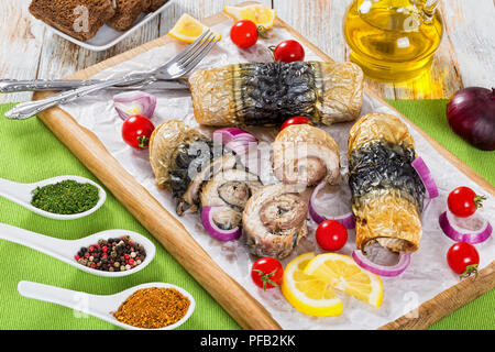 Gesunde gebackenes Filet von der Makrele in Brötchen, Tomaten und Zitronenscheiben auf weißem Pergamentpapier auf Schneidebrett mit Messer und Gabel, Gewürze, Roggenbrot Stockfoto