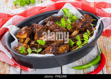 Leckere heiße klebrigen Ribs gewürzt mit würzigem Knoblauch Ingwer Barbecue Sauce und Koriander in ovalen Bräter auf weißen Planken mit Küche Tuch. gre Stockfoto