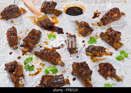 Leckere heiße klebrigen Ribs gewürzt mit pikanten Soja Knoblauch Ingwer Barbecue Sauce auf Pergamentpapier mit Pinsel und Korianderblätter, auf Holz- Peeling Stockfoto