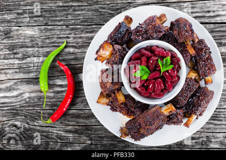 Gekochte rote Bohnen in der Schüssel durch gegrillte Rippen auf weiße Platte umgeben, zwei Chili auf alten Holzdielen, Ansicht von oben Stockfoto