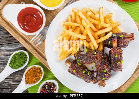 Gebratene klebrige Rippen bestreut mit grüne Zwiebeln mit Pommes frites auf weißem Teller auf Schneidebrett mit Senf und Tomatenmark in Soße Boote, auf die Tabelle m Stockfoto
