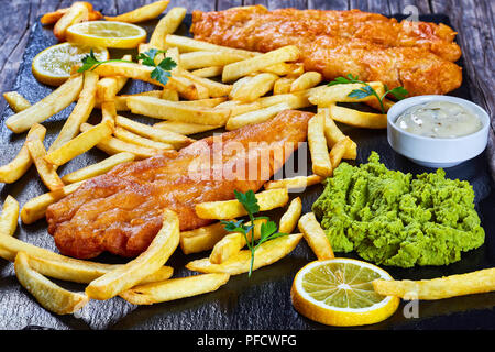 Köstlich knusprige Fisch und Chips auf schwarzem Schiefer Platte mit Sauce Tartare, matschig Erbse, Zitronenscheiben auf dunklem Hintergrund, Ansicht von oben, Nahaufnahme, Stockfoto