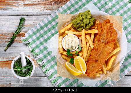 Knusprige Fisch und Chips - gebratener Kabeljau, Pommes frites, Zitronenscheiben, Remoulade und Erbsenpüree auf Platte auf Papier auf alten Holztisch mit Rosmarin in Mor Stockfoto