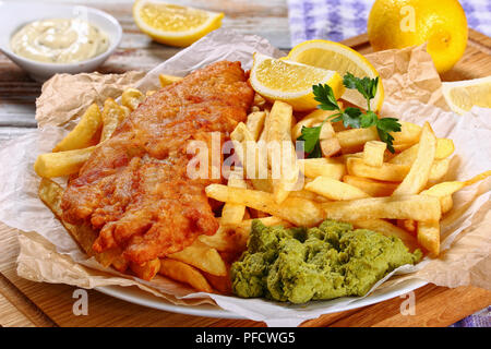 Knusprige Fisch und Chips - gebratener Kabeljau, Pommes frites, Zitronenscheiben, Remoulade und Erbsenpüree auf Papier auf Holz Schneidebrett, Zitrone und Küche Tuch Stockfoto