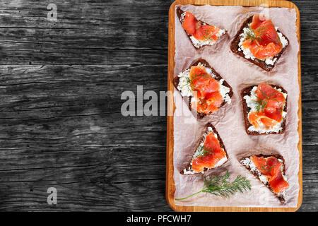 Leckeren Toast mit Frischkäse und Lachs auf Vollkorn Brot mit Sonnenblumenkernen auf Schneidebrett auf dunklen Holz- Tabelle, Ansicht von oben Stockfoto