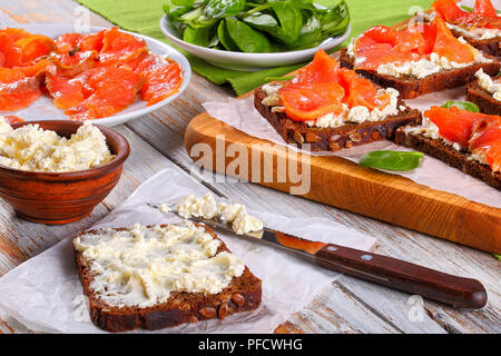 Köstliche Roggen Brot Toast mit Frischkäse erstickt und gekrönt mit Lachs Scheiben auf weißem Papier auf Schneidebrett. Schale mit frischem Spinat und Salat Stockfoto