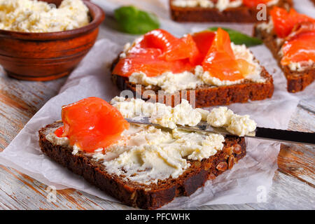 Roggen Brot Toast mit Frischkäse erstickt und gekrönt mit Lachs Scheiben, Messer und Schüssel mit Käse auf weißem Papier mit Spinatblättern, Ansicht von eine Stockfoto