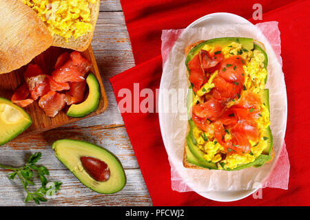 Lachs, Avocado Rührei ciabatta leckeren Sandwich mit Petersilie auf weißem Papier auf ovalen Teller bestreut, mit Zutaten auf hölzernen Tisch, Blick fr Stockfoto