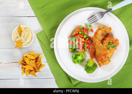 Teil der goldenen Teig frittierte Meer Fischfilets mit Petersilie dekoriert, auf weiße Platte mit einem Mix aus Dampf gekocht Gemüse serviert, getreidemehl Chips Stockfoto