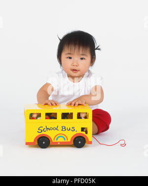 Baby Mädchen in roten Hosen sitzen auf dem Boden mit Schule des gelben Spielzeug bus' Figuren vor ihrer Durchführung, Vorderansicht. Stockfoto