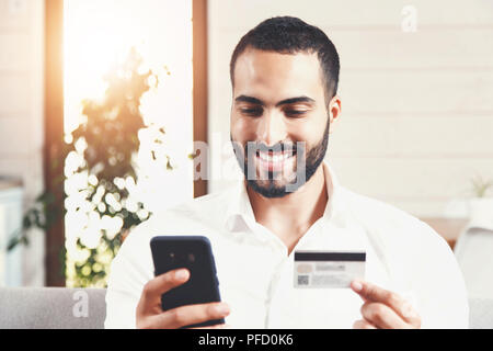 Gut aussehender bärtiger Mann im weißen Hemd auf dem Sofa sitzen, die online Kauf über das Smartphone mit der Kreditkarte Stockfoto