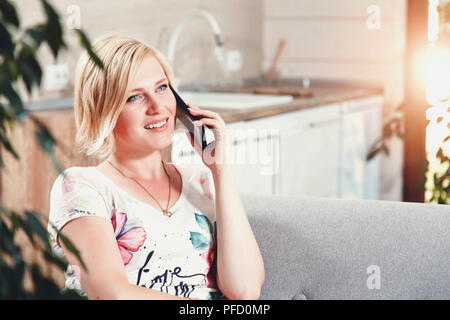 Hübsche Blondine lächelnde Frau auf dem Sofa sitzen und sprechen auf dem Smartphone in weiten studio Küche Stockfoto