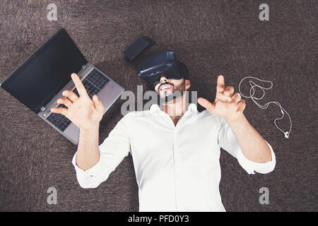 Bärtiger Mann mit Vr-set Brillen liegen auf dem Boden neben dem Laptop, Smartphone und Kopfhörer Stockfoto