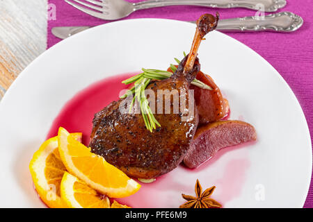 Gebratene Entenkeule in Rotwein mit Rosmarin, Orange und Apfel Scheiben auf einem weißen Teller auf ein Schneidbrett, Ansicht von oben, close-up Stockfoto