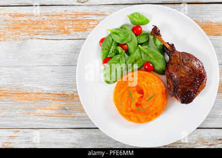 Gebratene Entenkeule in Rotwein mit Rosmarin, Orange und Apfel Scheiben auf weissem Teller mit Spinat und Kürbis porridge auf ein Schneidbrett, Ansicht von oben, Stockfoto