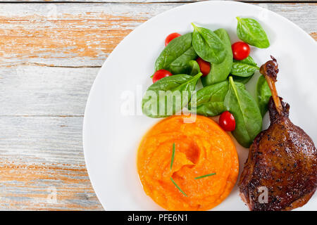 Gebratene Entenkeule in Rotwein mit Rosmarin, Orange und Apfel Scheiben auf weissem Teller mit Spinat und Kürbis porridge auf ein Schneidbrett, Ansicht von oben, Stockfoto