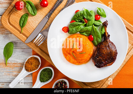 Gebratene Entenkeule mit Kartoffelpüree Kürbis, Spinat und Tomaten Salat auf einem weißen Teller auf Schneidebrett mit Gabel, Messer und Gewürze in Keramik Löffel, auf Rost Stockfoto