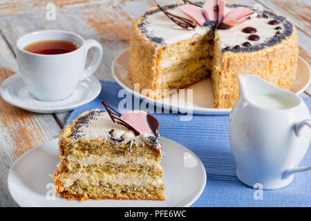 Biskuitteig mit Mohn, überlagert mit Creme mit ein ausgeschnittenes Stück auf weiße Platte close-up auf Tisch Matte mit Krug Milch, Tasse mit schwarzem Kaffee auf SAUC Stockfoto