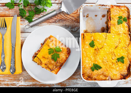 Teil der leckeren Moussaka auf Platte mit Messer und Gabel und in der auflaufform auf alten Holzbohlen, tradihional griechische Küche, authentische Rezept, Aussicht f Stockfoto