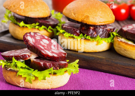 Köstliche Sandwiches mit Butter, Salat und Scheiben geräucherte Wurst in gebratene Brötchen auf dunklen Kammer auf Tisch Matte, Nahaufnahme, Ansicht von oben Stockfoto