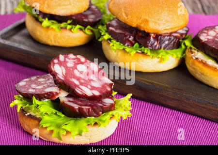 Köstliche Sandwiches mit Butter, Salat und Scheiben geräucherte Wurst in gebratene Brötchen auf dunklen Holzplatte auf Tisch Matte, Nahaufnahme, Ansicht von oben Stockfoto