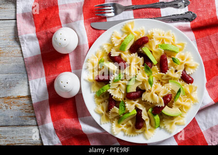Italienische Wurst Bow Tie Pasta warmer Salat mit Avocado Scheiben und Blätter Basilikum auf weißen Teller am Tisch Tuch mit Salz- und Pfefferstreuer, Gabel und kni Stockfoto