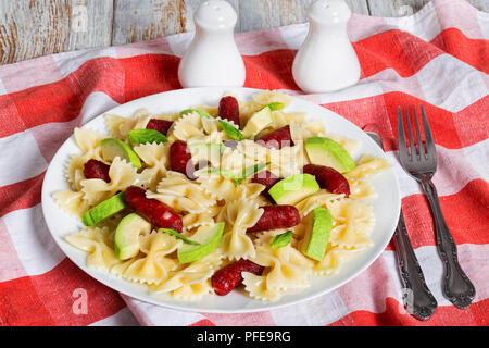 Italienische Wurst Bowtie Pasta warmer Salat mit Avocado Scheiben und Blätter Basilikum auf weißen Teller am Tisch Tuch mit Salz- und Pfefferstreuer, Gabel und knif Stockfoto