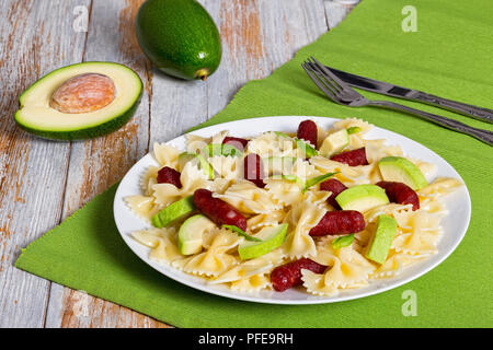 Mini Salami farfalle Pasta warmer Salat mit Avocado Scheiben und Blätter Basilikum auf weißen Teller am Tisch mit Messer und Gabel, die Hälfte der reife Avocado auf ol Stockfoto