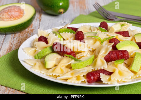Mini Salami farfalle Pasta warmer Salat mit Avocado Scheiben und Blätter Basilikum auf weißen Teller am Tisch mit Messer und Gabel, ganze und halbe reifer Avo Stockfoto