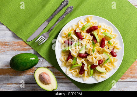 Mini salami Bowtie Pasta warmer Salat mit Avocado Scheiben und Blätter Basilikum auf weißen Teller am Tisch mit Messer und Gabel, die Hälfte der reife Avocado auf alten Stockfoto