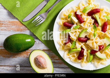 Mini Salami farfalle Pasta warmer Salat mit Avocado Scheiben und Blätter Basilikum auf weißen Teller am Tisch mit Messer und Gabel, ganze und halbe reifer Avo Stockfoto